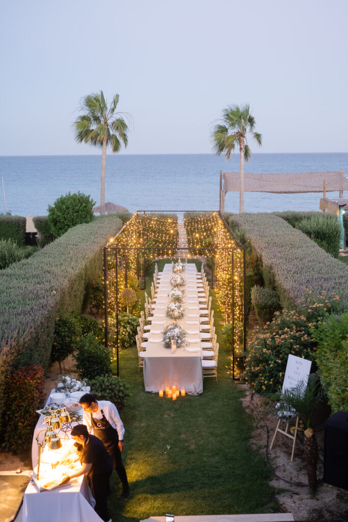 Magnificent outdoor wedding reception at Atlantis Gardens in Mazotos, featuring a long banquet table beneath a spectacular fairy light tunnel installation. The garden venue is styled with white floral centerpieces, gold-rimmed glassware, and elegant lighting, offering a luxurious destination wedding experience in Cyprus.