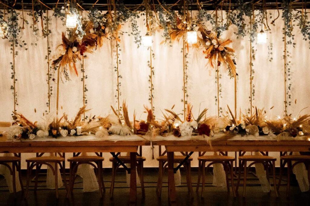 Warm and intimate wedding decor at Legacy venue featuring long wooden banquet tables and rustic cross back chairs. The backdrop is styled with hanging greenery vines, fresh flower arrangements in autumnal tones, and romantic pendant lighting, showcasing a cozy yet luxurious reception in Cyprus.