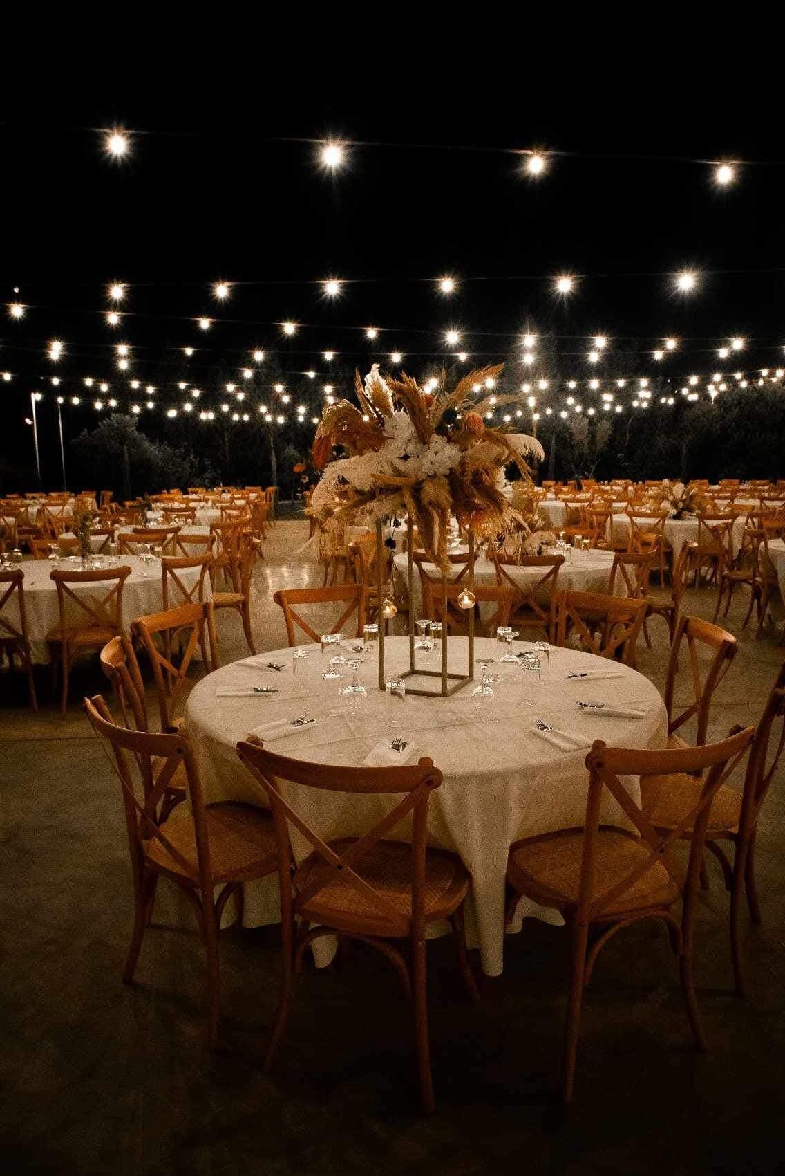 Atmospheric rustic wedding venue decor at Legacy venue, featuring round tables with white textured linens and wooden crossback chairs. The outdoor reception is illuminated by a massive fairy light canopy and features tall boho centerpieces with pampas grass, showcasing expert event lighting and rental styling in Cyprus.