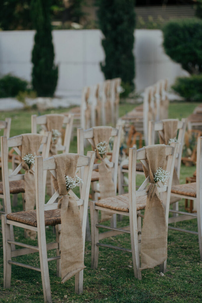 Outdoor rustic ceremony chair decor featuring white-washed wooden cross-back chairs with woven seats, adorned with burlap sashes and delicate baby's breath flowers. Professional rustic wedding furniture and decoration rentals in Cyprus for garden and beach ceremonies.