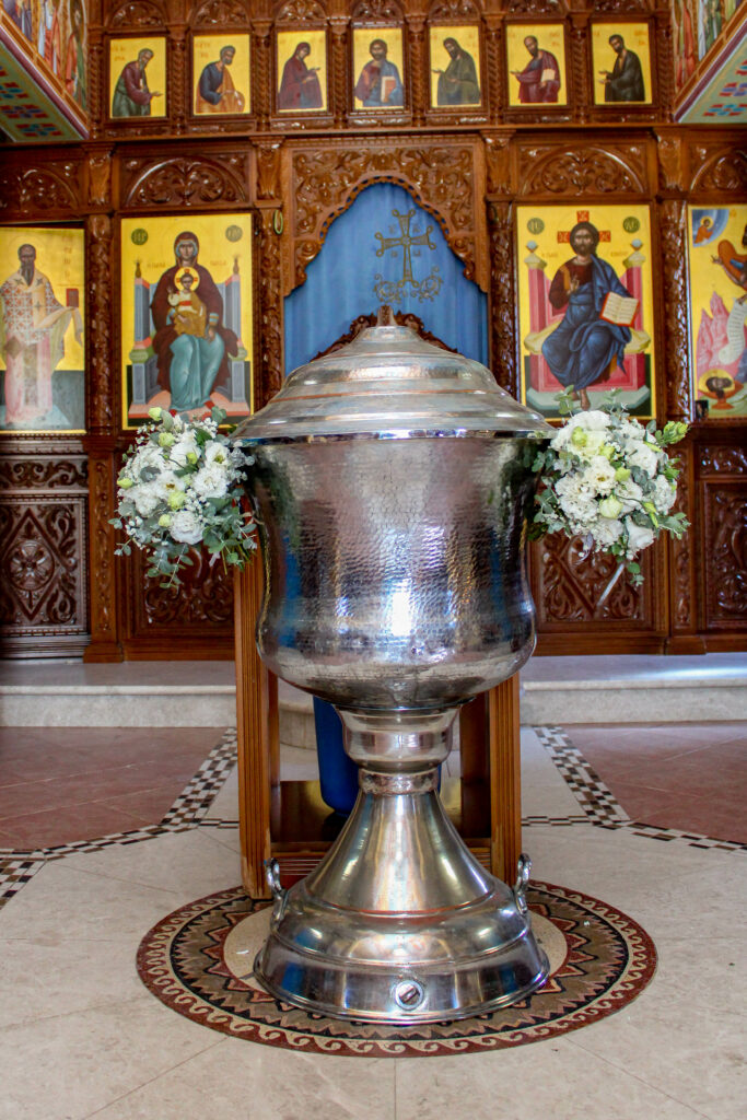 Traditional Orthodox baptismal font decoration in a Cypriot church, featuring elegant floral bouquets of white lisianthus, roses, and silver-green eucalyptus. The hammered silver Kolymbithra is adorned with symmetrical 'bourboula' floral arrangements, capturing the timeless and heartfelt essence of a Greek Orthodox christening in Cyprus