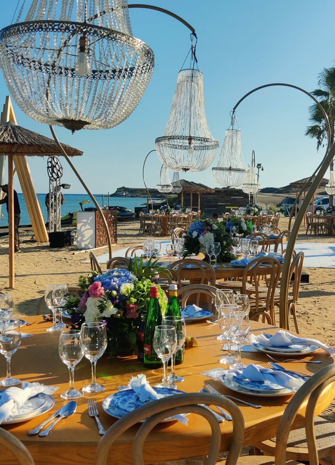 Luxury beach wedding reception in Cyprus featuring stunning crystal chandeliers hanging over circular wooden tables on the sand. This coastal setup includes vibrant blue and pink fresh flower arrangements, elegant crossback chairs, and a clear sea view, perfect for a high-end beachfront wedding in Ayia Napa or Protaras.