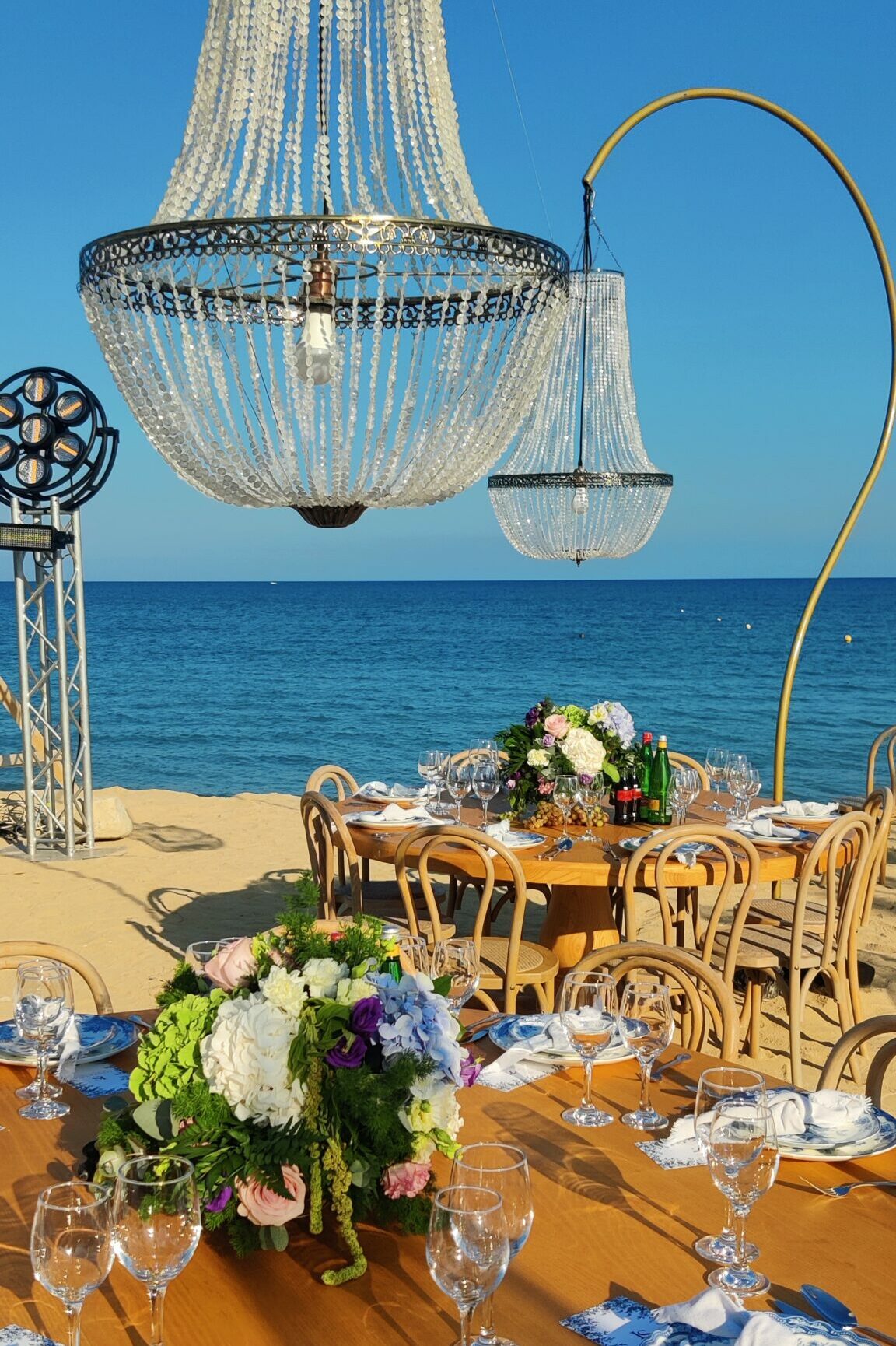 A high-end, bespoke beach event dining set-up on a Cyprus beach, featuring elegant blue and white pattern tableware, large floral centerpieces, and grand crystal chandelier rentals suspended overhead, with views of the Mediterranean Sea under a clear blue sky, demonstrating professional beach decoration rentals in Cyprus.