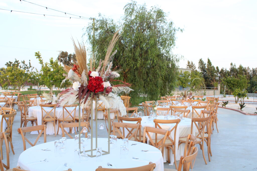 Outdoor boho wedding table decor in Cyprus featuring a tall gold stand centerpiece with vibrant red hydrangeas, pampas grass, and dried foliage. The alfresco reception setup at a rustic venue includes wooden crossback chairs and white linens, perfect for a modern boho-themed garden wedding