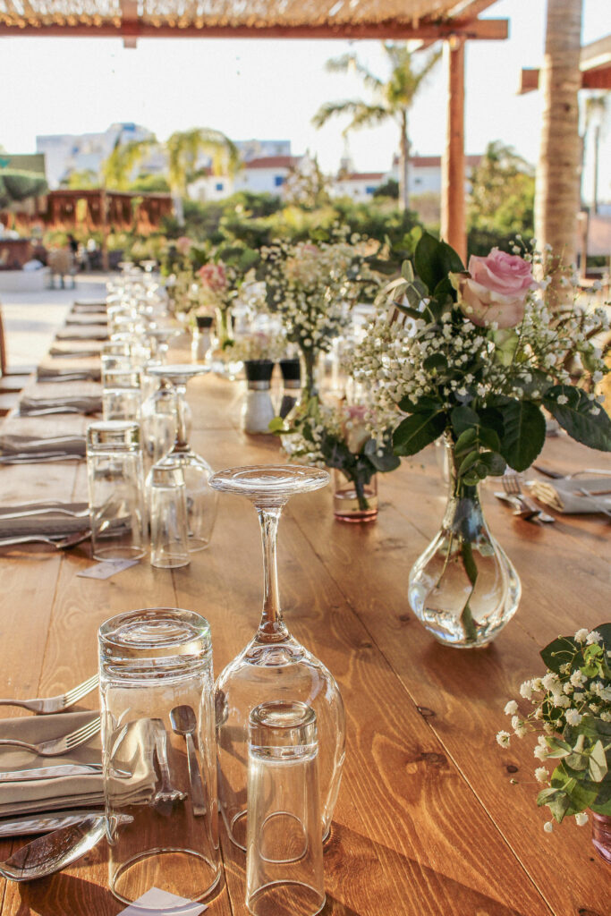 Elegant wedding reception at Cavo Zoe Seaside Hotel in Protaras, featuring long rustic wooden banquet tables and delicate fresh flower centerpieces. The seaside venue styling includes romantic pink roses, baby's breath in glass bud vases, and premium glassware, perfectly capturing the boho-chic Mediterranean wedding aesthetic.