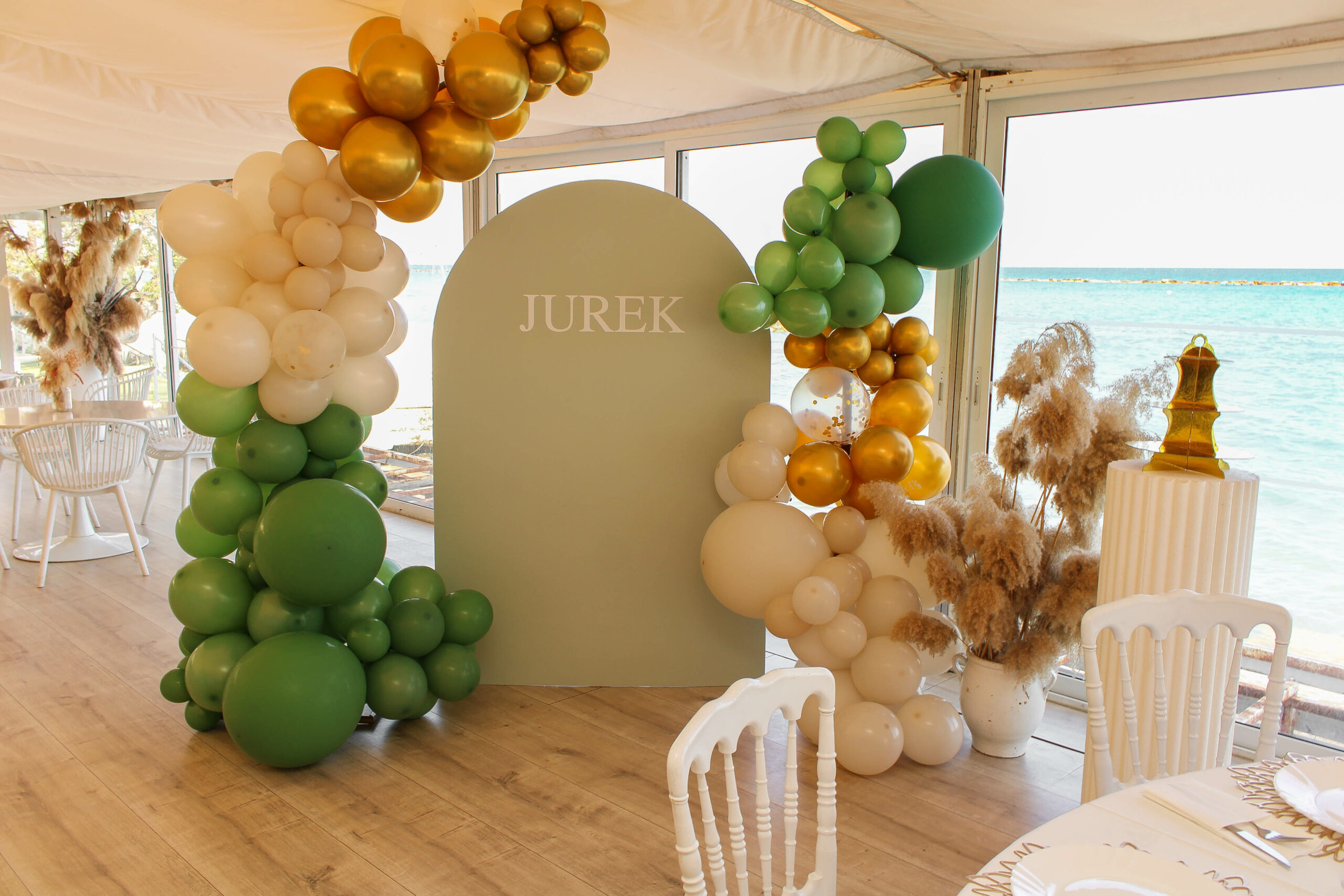 Elegant, sea-view christening decor at Nautica Larnaca, featuring a matte sage green arch with the personalized name 'JUREK'. The modern setup is framed by organic balloons in olive, sand, and gold, accessorized with tall pampas grass arrangements, perfect for a high-end baptism in Larnaca, Cyprus