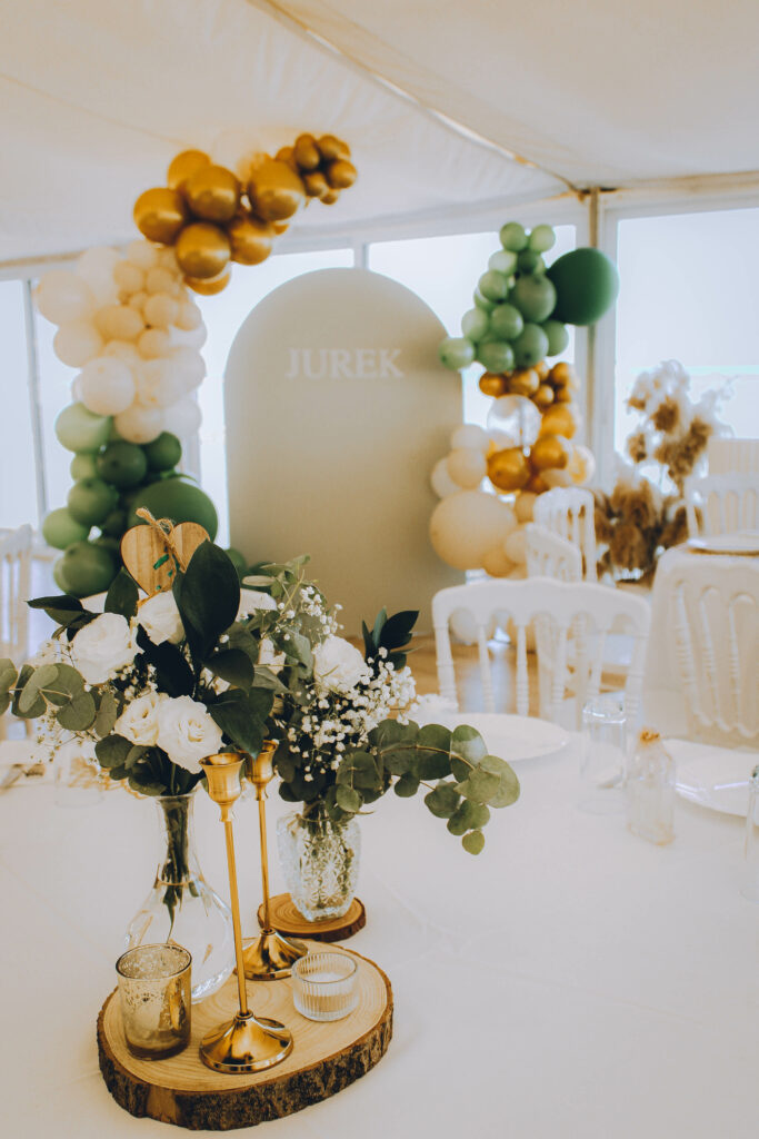 Bespoke sage green and gold table centerpiece for a luxury baptism reception in Cyprus. The setup features a rustic wood slice base, gold mercury glass candle holders, and delicate floral vases with white roses, baby's breath, and eucalyptus, perfectly coordinating with a modern olive green balloon backdrop.