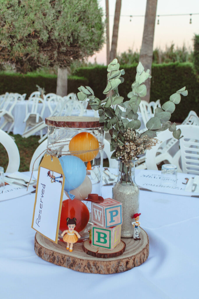 Bespoke outdoor party table decor in Cyprus featuring a Toy Story themed centerpiece on a white linen table. The setup includes a rustic wood slice base, a sand-filled glass bottle with fresh eucalyptus, and vintage alphabet blocks (P, B, L) paired with character figurines, creating a sophisticated and heartfelt garden celebration atmosphere.