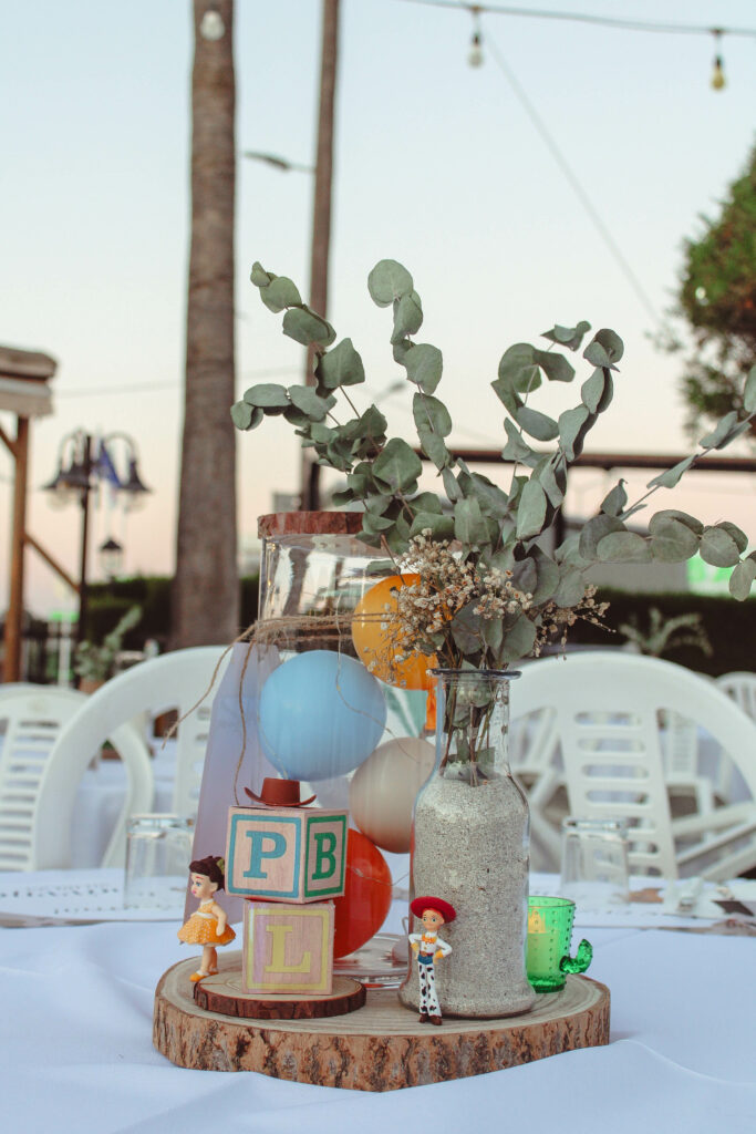 Custom Toy Story themed table centerpiece for a children's party, featuring a rustic wood slice base and a glass cylinder filled with colorful mini balloons. The nostalgic design is accented with alphabet toy blocks, vintage-style character figurines, and a sprig of fresh eucalyptus in a sand-filled bottle, ideal for a heartfelt family birthday in Cyprus.