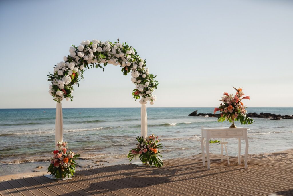 An elegant outdoor wedding ceremony setup on a sun-drenched European beach, featuring a white floral arch overlooking the turquoise Mediterranean Sea. The scene is set on a wooden deck with a vintage white signing table and lush tropical flower arrangements.