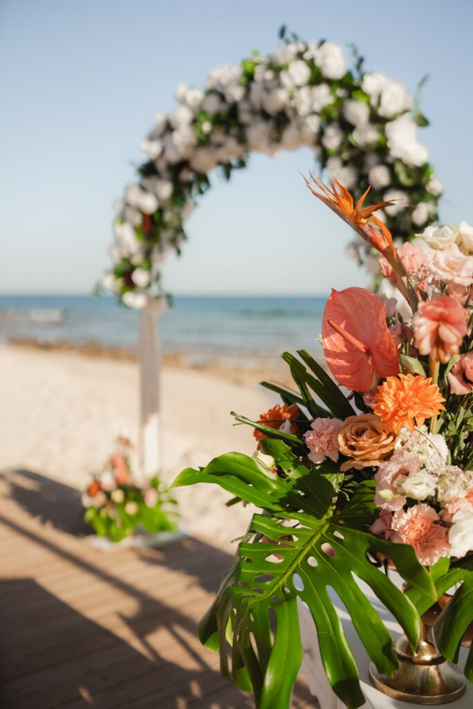 Elegant tropical beach wedding décor at Sirens beach wedding venue, featuring a floral ceremony arch with white roses and greenery, coral and peach floral arrangements with monstera leaves, and stylish seaside aisle setup. Romantic coastal wedding decoration showcasing premium beach wedding styling and decoration rental services in Cyprus