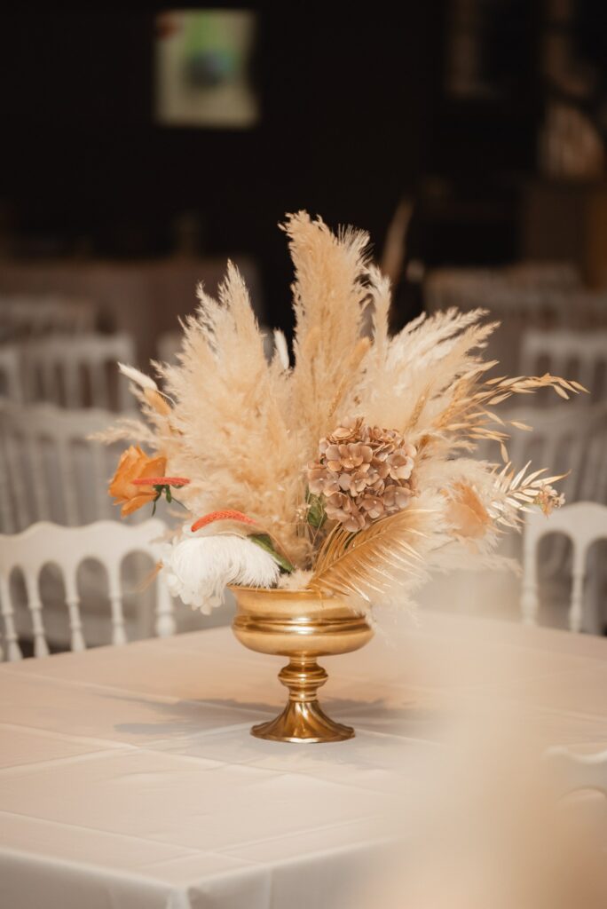 Elegant boho wedding centerpiece in a gold pedestal bowl, featuring a mix of fresh flowers and dried pampas grass. This luxurious Cyprus venue decoration includes terracotta-toned roses and textured grasses, perfectly suited for a modern boho-themed wedding reception with white Napoleon chairs.