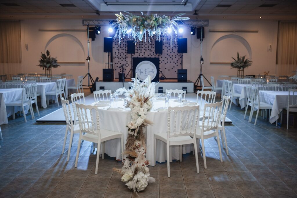 Modern indoor wedding reception rentals in Cyprus featuring a luxurious hanging floral installation above the dance floor. The setup includes elegant white Napoleon chairs, crisp white tablescapes, and a pampas grass floral runner cascading from the head table, creating a sophisticated all-white wedding theme for a luxury event.