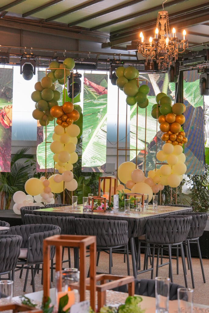 Tropical event decor rentals at Velissima Club featuring green and gold balloon installations, wood centerpieces, and chic woven seating for premium parties in Cyprus.