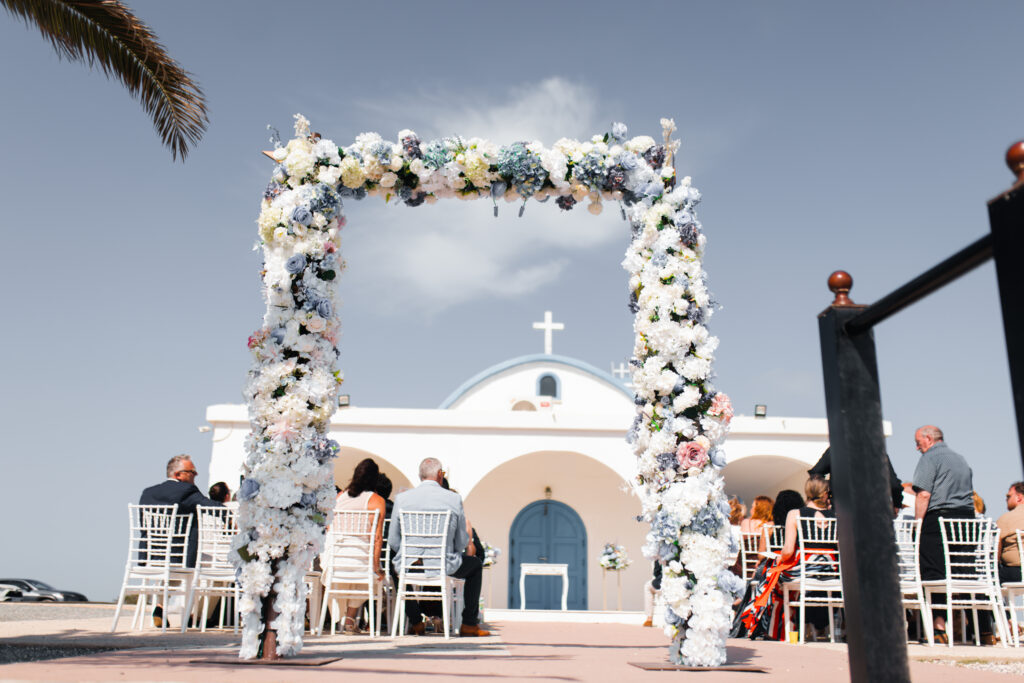 Outdoor wedding ceremony in Cyprus featuring a large floral archway with white and light blue flowers. Guests are seated in white chivari chairs facing a traditional Greek Orthodox church with a blue door and dome. Ideal for Cyprus church wedding decor rentals and destination wedding styling.