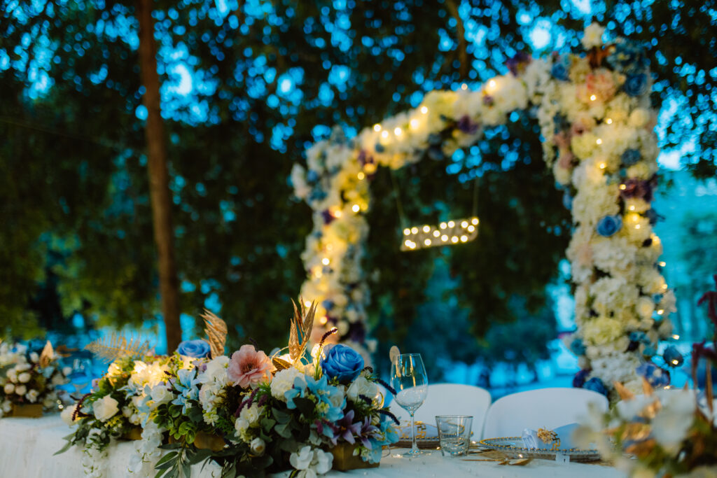 Romantic outdoor wedding reception at night featuring a massive floral arch with fairy lights. The head table is adorned with vibrant blue and pink fresh flower arrangements, gold cutlery, and elegant glassware, showcasing premium wedding venue decoration in Cyprus for an enchanted garden theme.