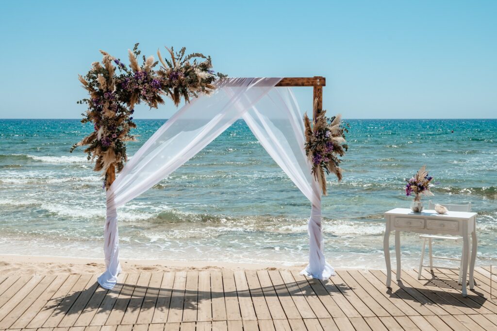 Square wooden ceremony arch with white fabric, purple and boho decor and white ceremony table styled by professional decoration company in Cyprus