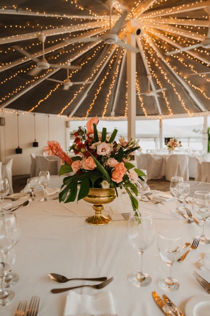 Vibrant wedding reception at Nissi Beach Hotel Rondavel, featuring a tropical floral centerpiece in a gold pedestal bowl with monstera leaves and pink roses. The circular venue is illuminated by a stunning ceiling fairy light canopy, showcasing luxury wedding decorations in Ayia Napa for a glamorous seaside celebration.