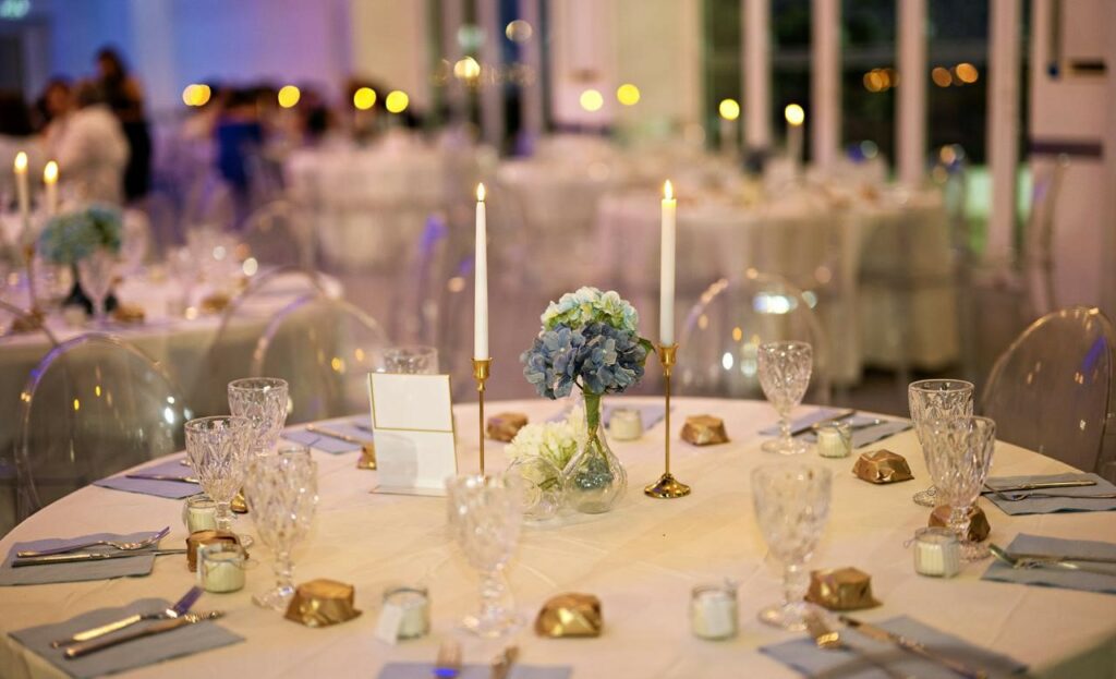 Elegant evening wedding table styling in Cyprus featuring dusty blue hydrangea centerpieces and tall gold taper candle holders. The sophisticated tablescape includes crystal-cut glassware, ghost chairs, and fresh flower arrangements, creating a romantic candlelit ambiance for a modern luxury wedding reception.