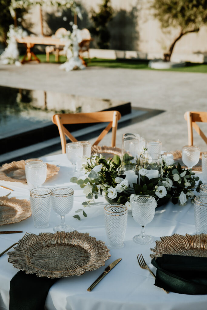 Luxury wedding tablescape at Legacy venue in Cyprus, featuring textured gold charger plates and crystal glassware. This poolside reception setup includes a lush white rose and eucalyptus centerpiece with floating candles, styled with wooden crossback chairs for a sophisticated modern destination wedding.