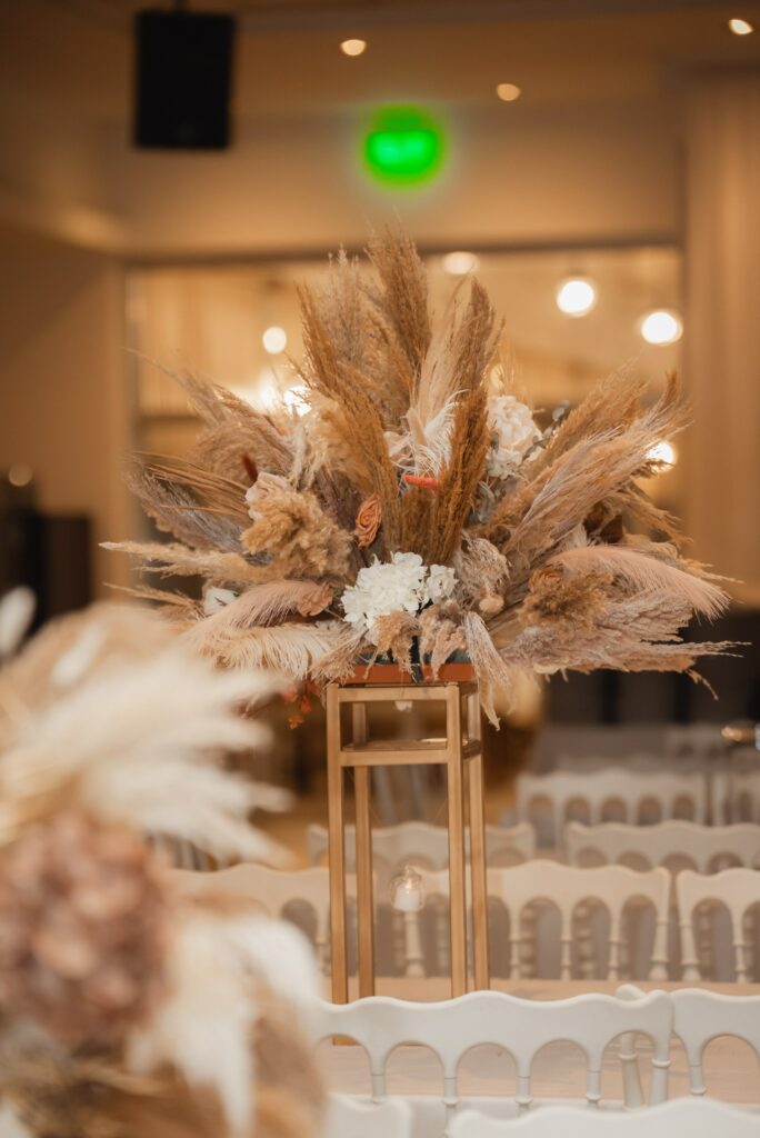 Grand indoor wedding at Laxia, Cyprus, featuring a spectacular tall boho centerpiece on a gold pedestal. This large-scale venue decoration includes a massive arrangement of dried pampas grass and white hydrangeas, styled with white Napoleon chairs to create a luxurious and airy feel for big wedding receptions.