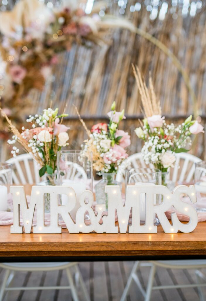Close-up of a rustic wedding head table in Cyprus featuring a lighted 'Mr & Mrs' wooden sign and delicate fresh flower arrangements in glass bud vases. The romantic setup includes soft pink ranunculus, white roses, and dried grass accents, perfectly styled for an alfresco destination wedding reception.