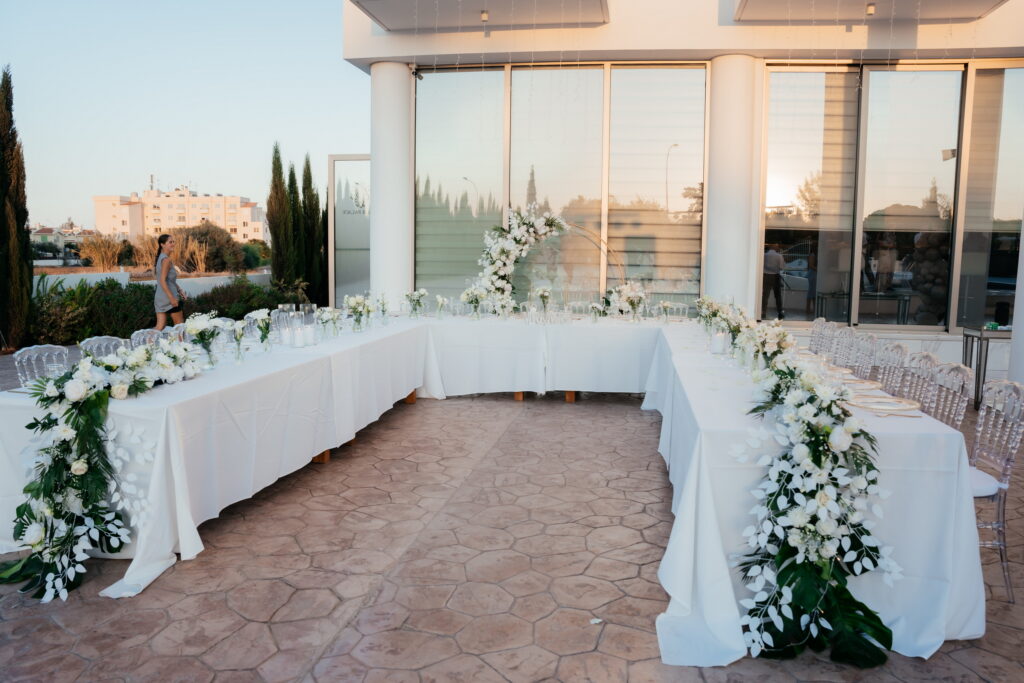 Luxury private villa wedding decor at Maxor Palace, Cyprus, featuring a grand U-shaped head table with crisp white linens. This sophisticated outdoor setup includes a cascading white floral runner, clear Napoleon ghost chairs, and a minimalist gold circular arch, showcasing premium wedding venue styling for exclusive estate celebrations.