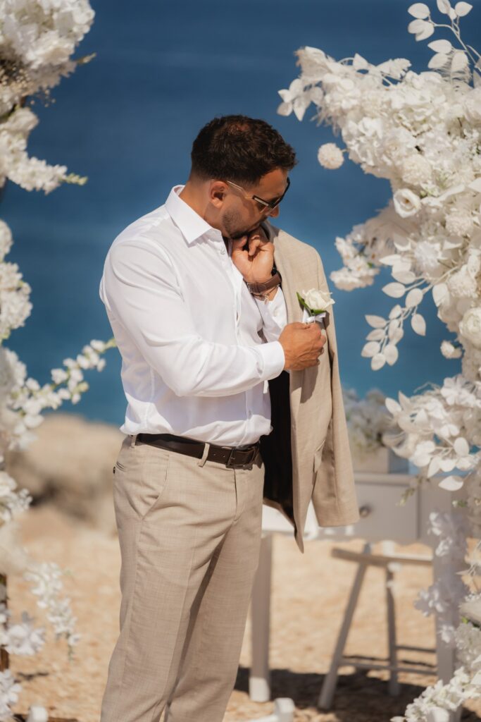 Cyprus wedding photoshoot groom in a beige suit in front of white floral arch at Blue lagoon in Cyprus