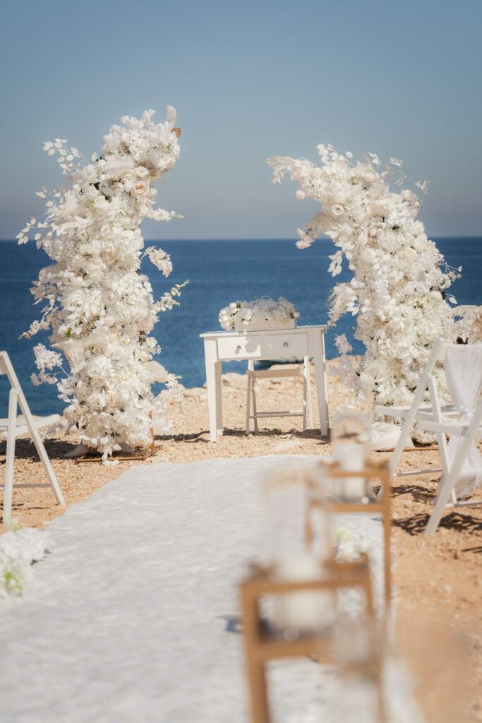 white half moon wedding arch insiration Luxury white floral arch rentals for a beachfront wedding ceremony in Cyprus, featuring elegant all-white floral installations, a white aisle runner, and romantic coastal wedding decor overlooking the Mediterranean Sea.