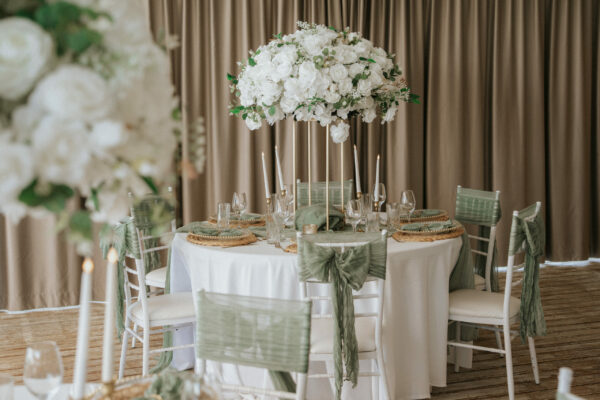 Di Capilla wedding large silk white floral wedding table centerpiece with white chivari chairs, green bows and luxury brown backdrop as a part of a wedding set up